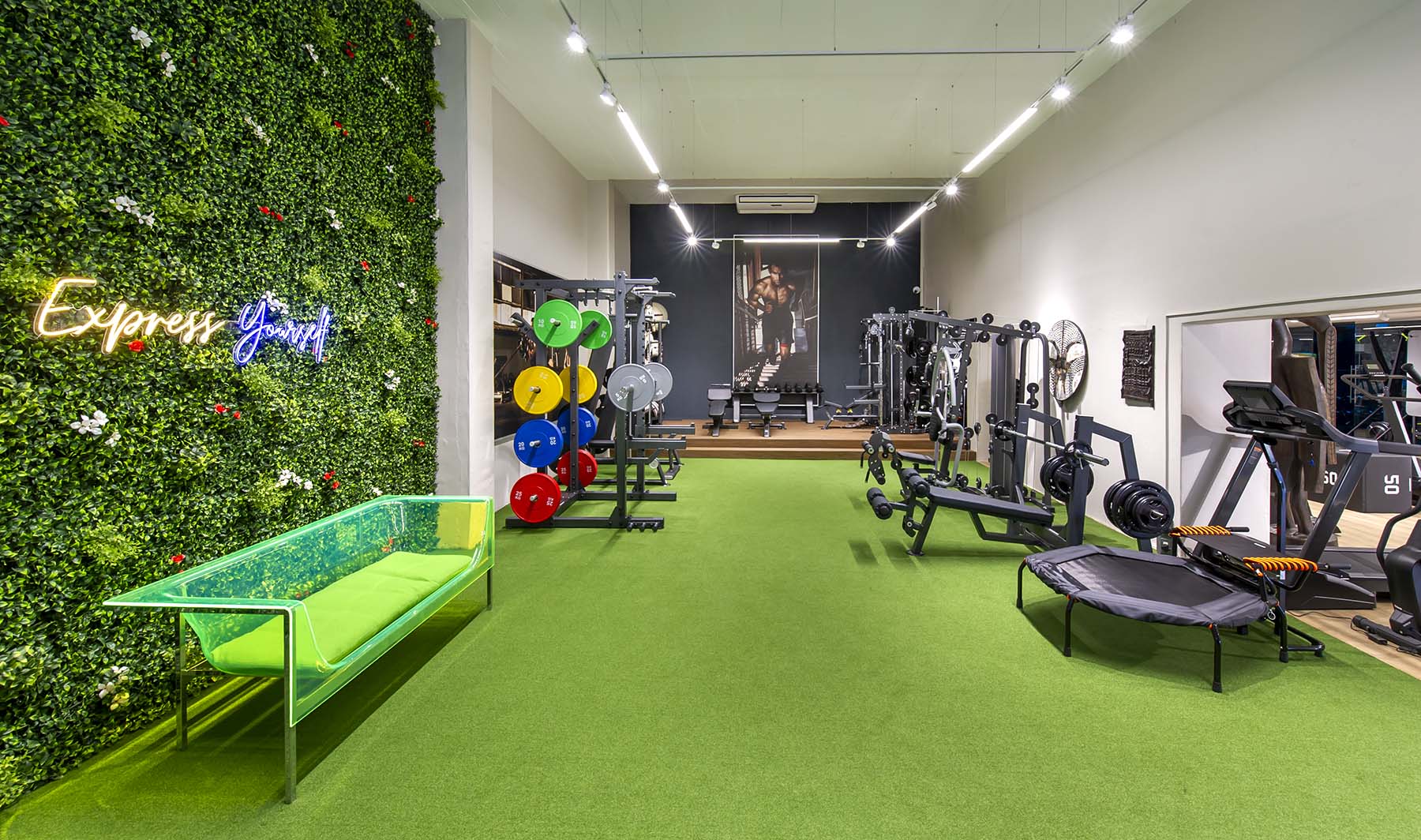 A modern Express Fitness Urban showroom with a vibrant green vertical garden wall and a neon sign. The floor is green artificial turf, and the space is filled with strength training and cardio equipment.