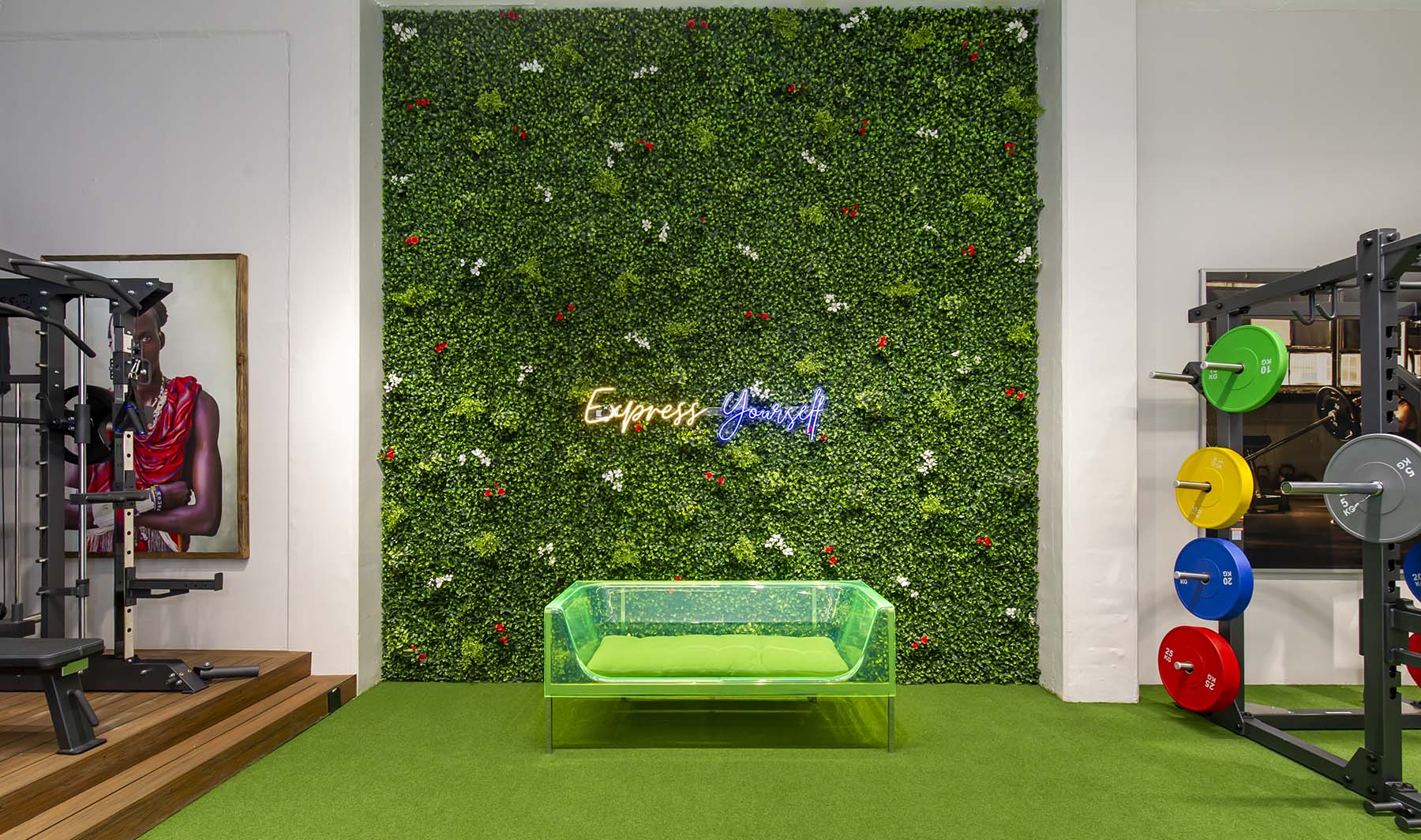 A modern Express Fitness Urban showroom with a large green vertical garden wall and a neon sign. A green bench is in front, and a framed painting of a Maasai warrior hangs on the left wall.