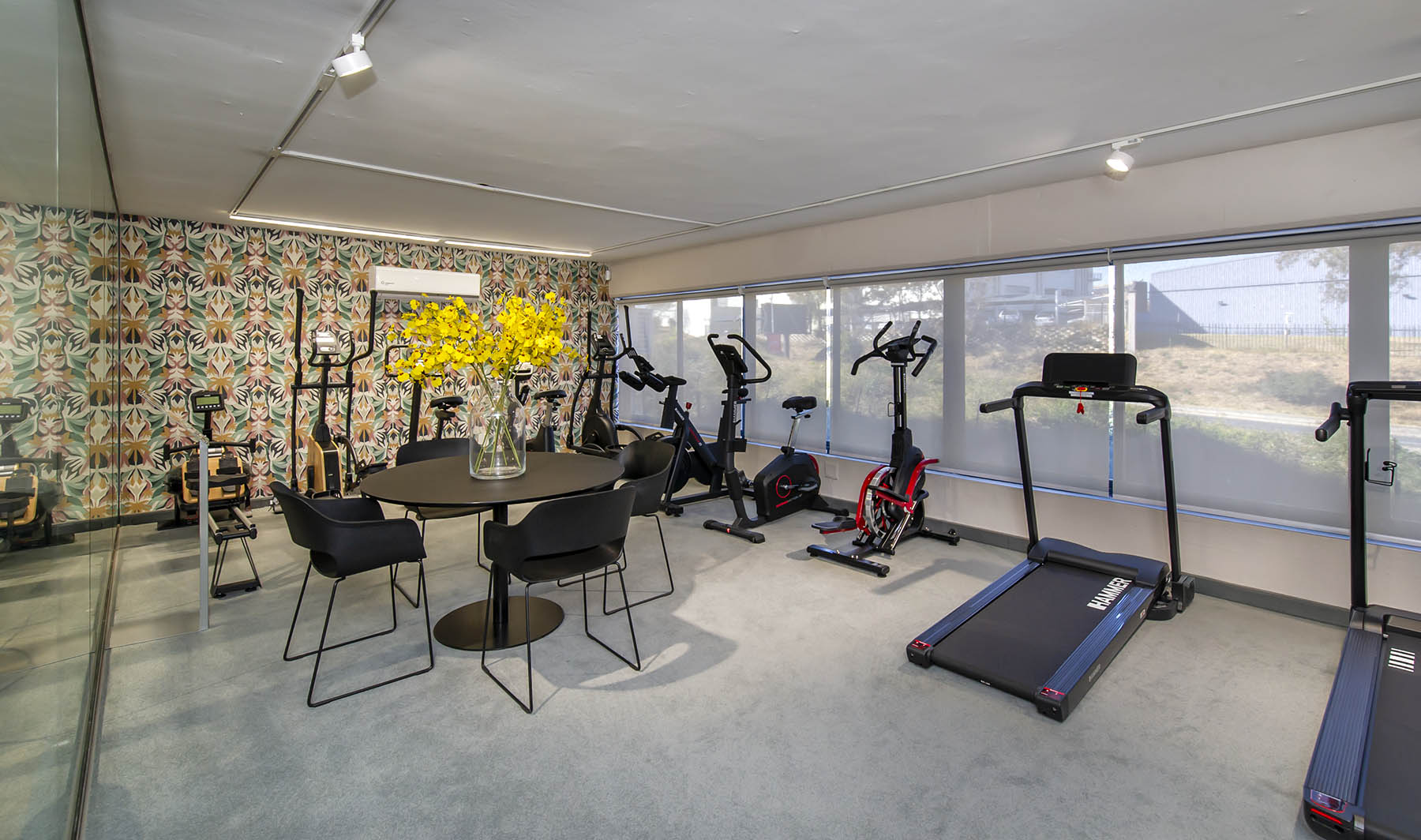 A bright and stylish meeting area within the Express Fitness Urban showroom in Sandton, Gauteng, featuring patterned wallpaper, large windows, and a round consultation table with chairs. Various cardio machines including treadmills, ellipticals, and exercise bikes are also displayed in the background, showcasing a range of home gym equipment.