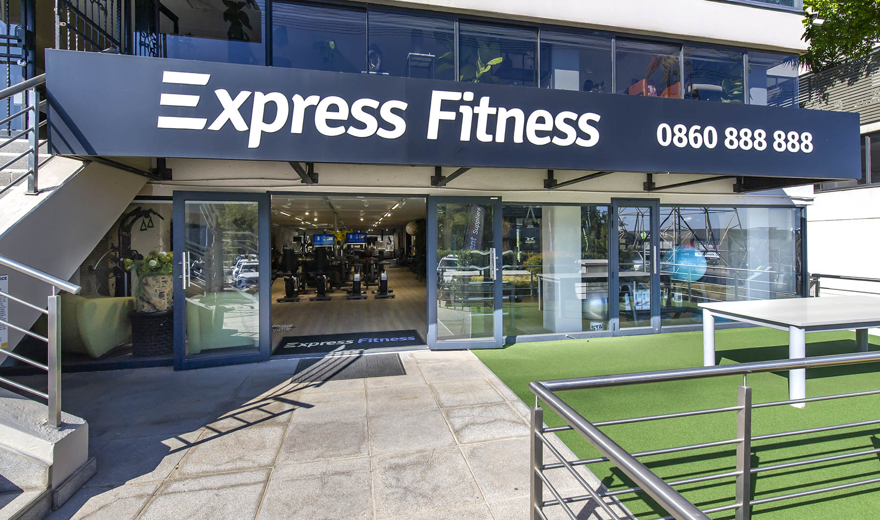 The exterior of Express Fitness Urban showroom in Sandton. The building has a prominent sign, large glass windows, and open doors leading to the gym equipment display. An outdoor patio with artificial turf is visible on the right.